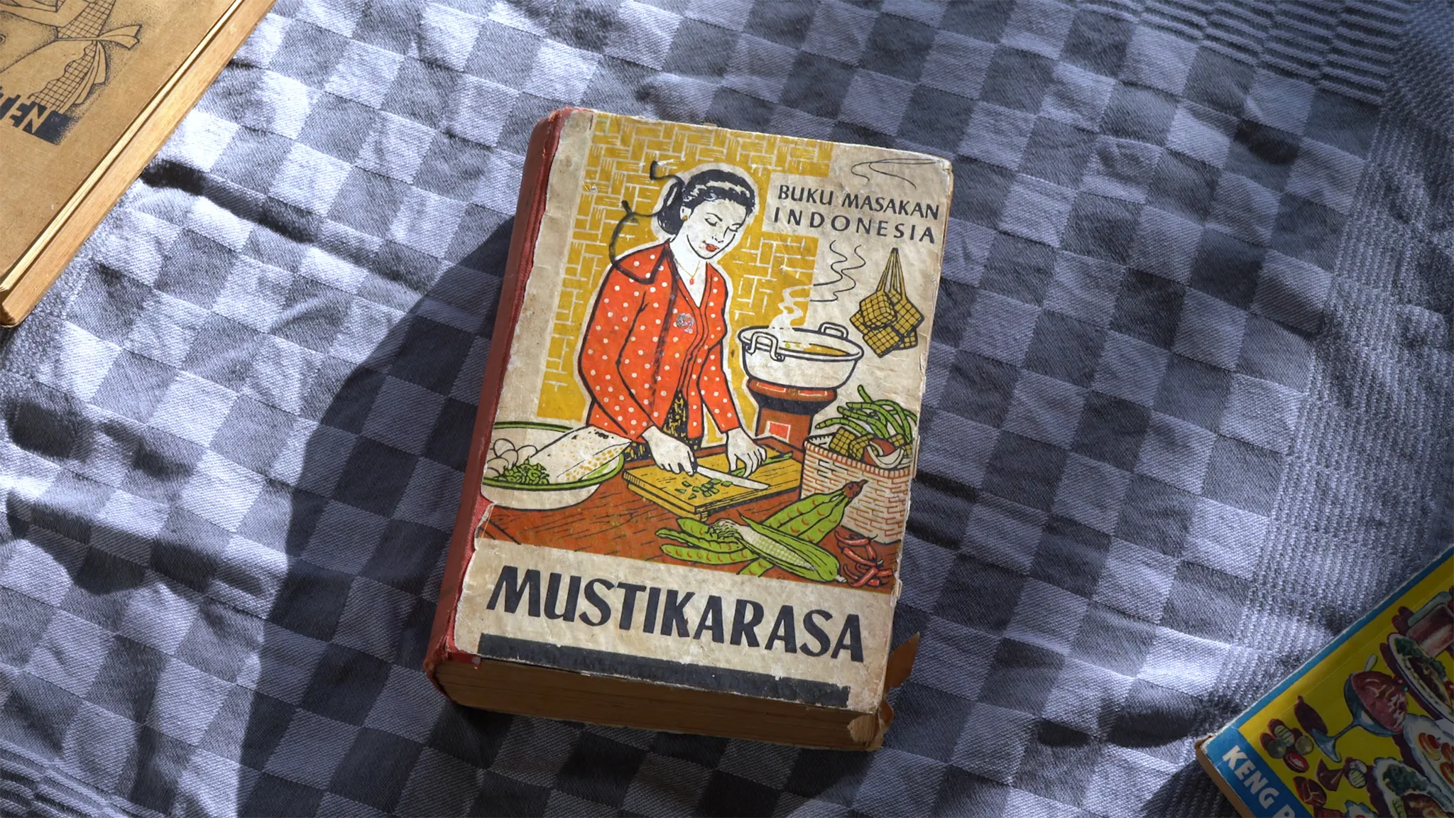 Een oud Indonesisch kookboek met een getekende omslag op een blauwe keukenhanddoek met vierkantjes.