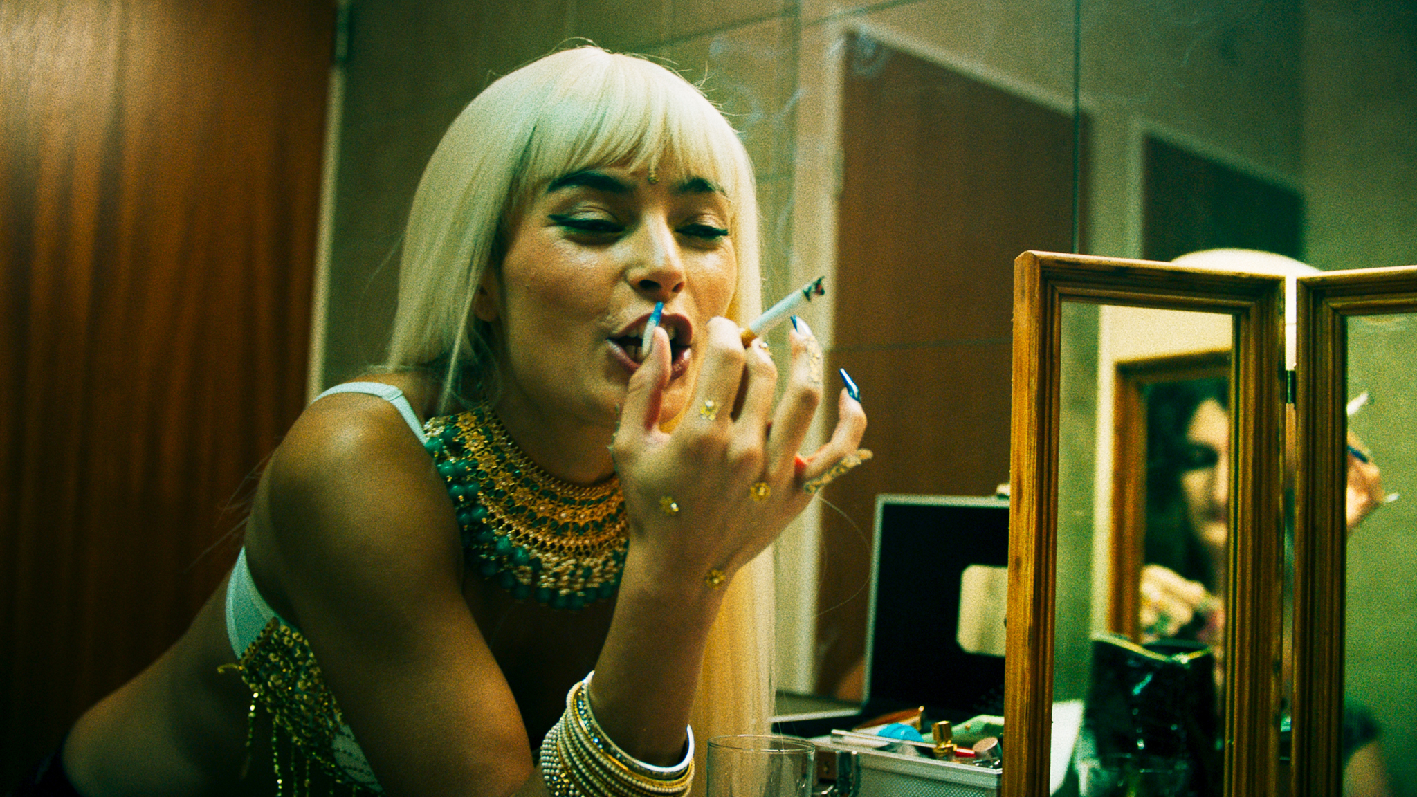 Een blonde persoon leunt op een meubel terwijl ze lachend een cigarette rookt.
